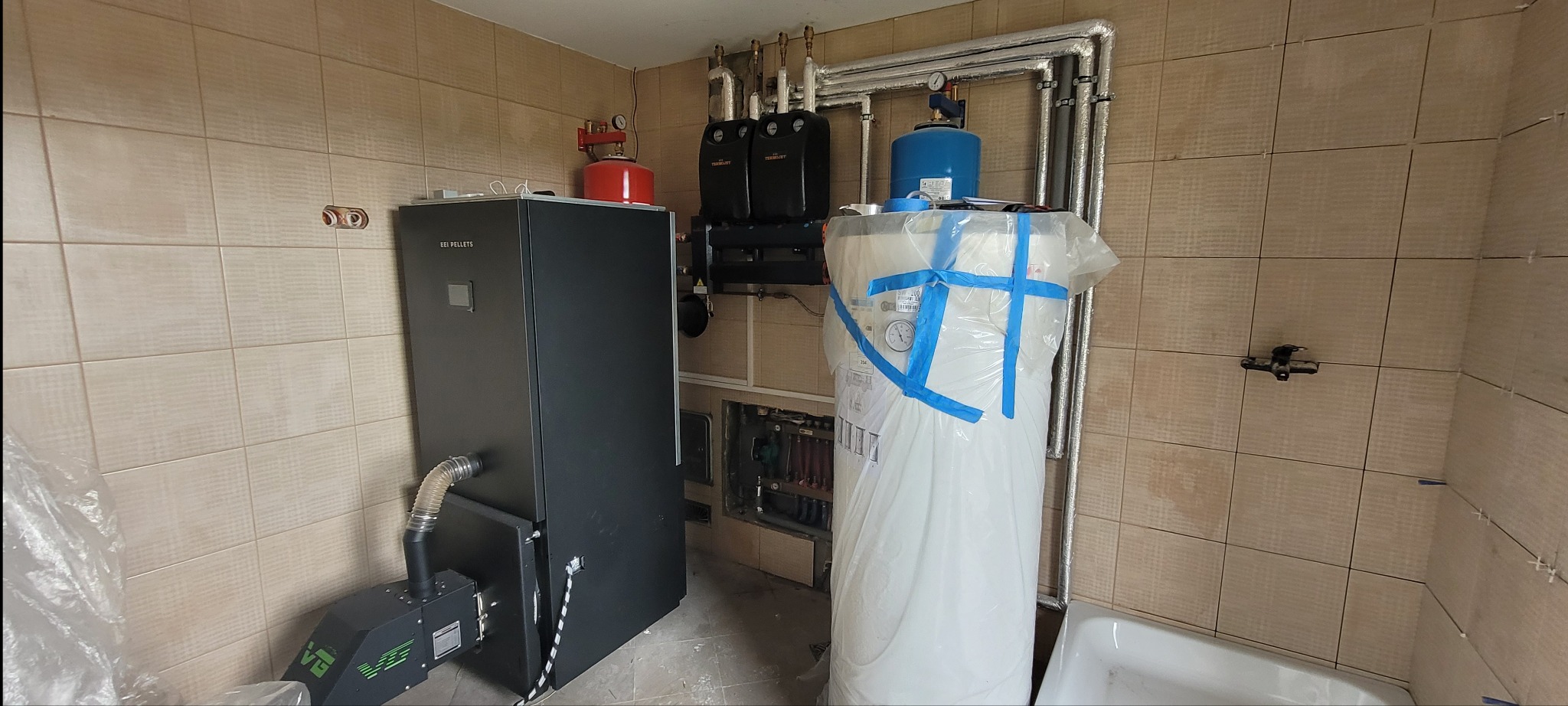 Kotłownia w trakcie montażu: piec na pellet, zbiornik buforowy zabezpieczony folią, instalacja grzewcza z rurami w izolacji, płytki na ścianach z widocznymi krzyżykami dystansowymi.