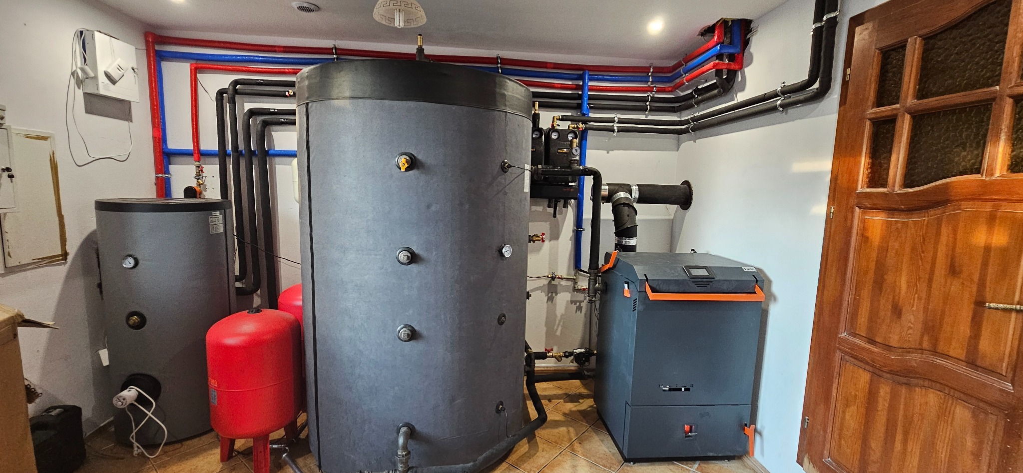 Kotłownia z dwoma zbiornikami akumulacyjnymi, kotłem grzewczym i instalacją z rur w kolorach czerwonym, niebieskim i czarnym, widoczna drewniana stolarka drzwiowa.