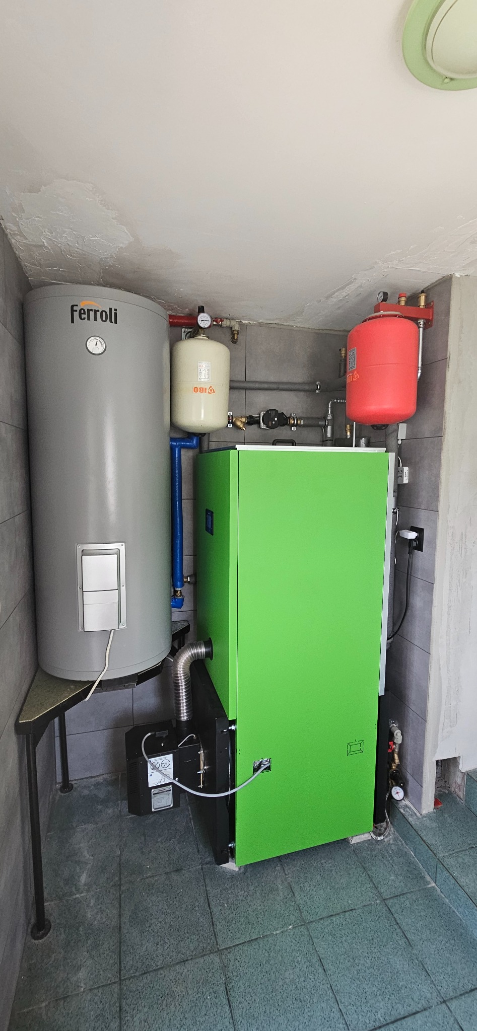 Zestaw urządzeń grzewczych: zielony piec, szary bojler Ferroli na metalowym stelażu, biały i czerwony zbiornik wyrównawczy, zamontowane w kotłowni z szarymi płytkami ściennymi i podłogowymi.