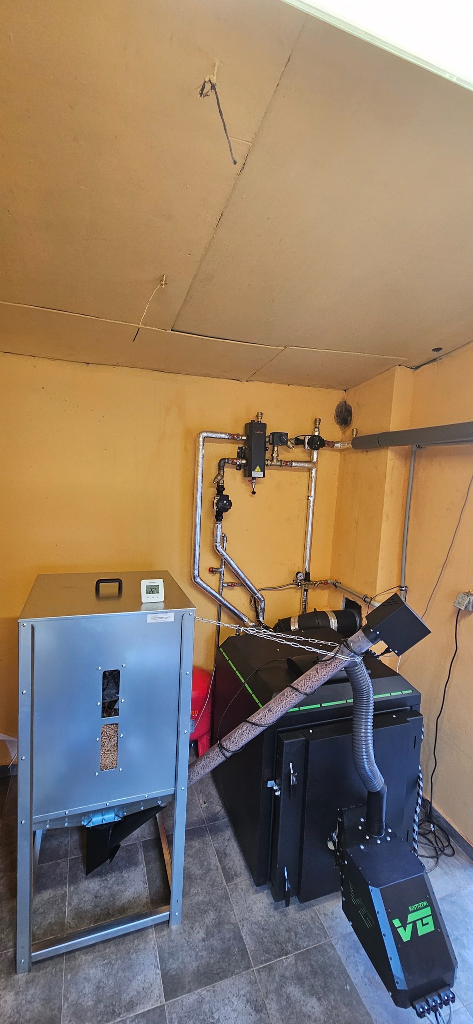 Kotłownia z podajnikiem na pellet i rurami instalacji grzewczej na żółtej ścianie, widoczne elementy automatyki i zbiornik na pellet.