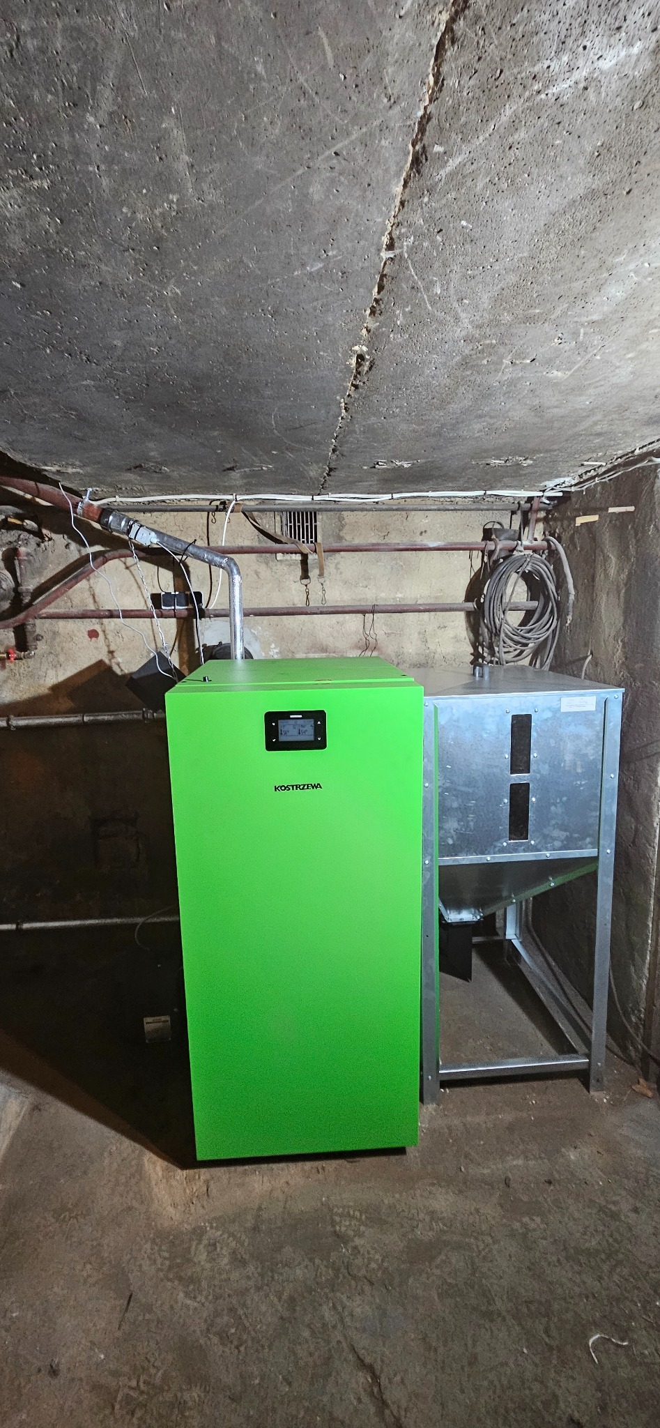 Jasnozielony piec Kostrzewa z podajnikiem w pomieszczeniu z surowym betonowym sufitem i widocznymi instalacjami.
