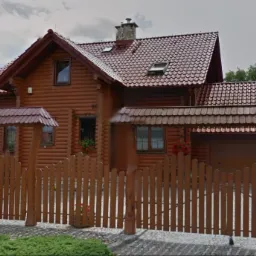 Urokliwy, drewniany dom z czerwonym dachem i ozdobnym ogrodzeniem w rustykalnym stylu. Widoczne kwiaty w oknach i zadbana zieleń.