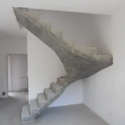 Surowe, betonowe schody w jasnym wnętrzu, widoczne z dołu pod kątem. Schody łączą dwa poziomy, a ich konstrukcja jest widoczna w stanie surowym przed wykończeniem.