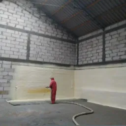 Pracownik w czerwonym kombinezonie natryskuje izolację na ścianę z bloczków wewnątrz budynku z blachą falistą na dachu.