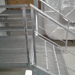 Metalowe schody kratowe z poręczami w surowym wnętrzu, widoczne elementy instalacji wentylacyjnej w tle.