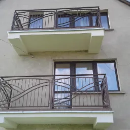 Dwa balkony z metalowymi balustradami o falistym wzorze na tle szarej elewacji budynku z oknami i anteną satelitarną.
