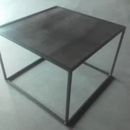 Stolik kawowy o minimalistycznej, surowej konstrukcji z metalowych profili kwadratowych i blatu z ciemnej blachy, ustawiony na szarym, betonowym podłożu.