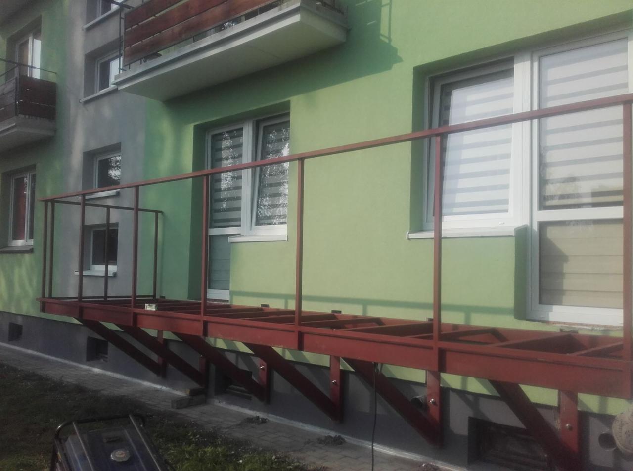 Konstrukcja stalowa balkonu w trakcie montażu, przymocowana do ściany budynku pomalowanej na zielono i szaro, widoczne okna z żaluzjami.