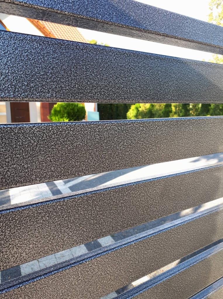 Fragment metalowego ogrodzenia panelowego o szorstkiej, ciemnoszarej fakturze, z widokiem na dom i zieleń w tle.