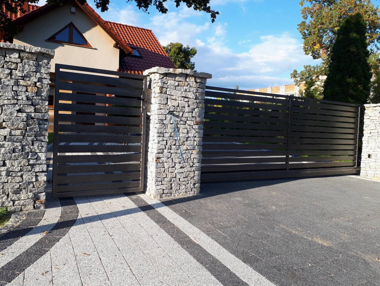 Nowoczesna, pozioma brama wjazdowa z szarego metalu, osadzona pomiędzy kamiennymi słupkami, na tle domu z czerwoną dachówką i ozdobną kostką brukową.