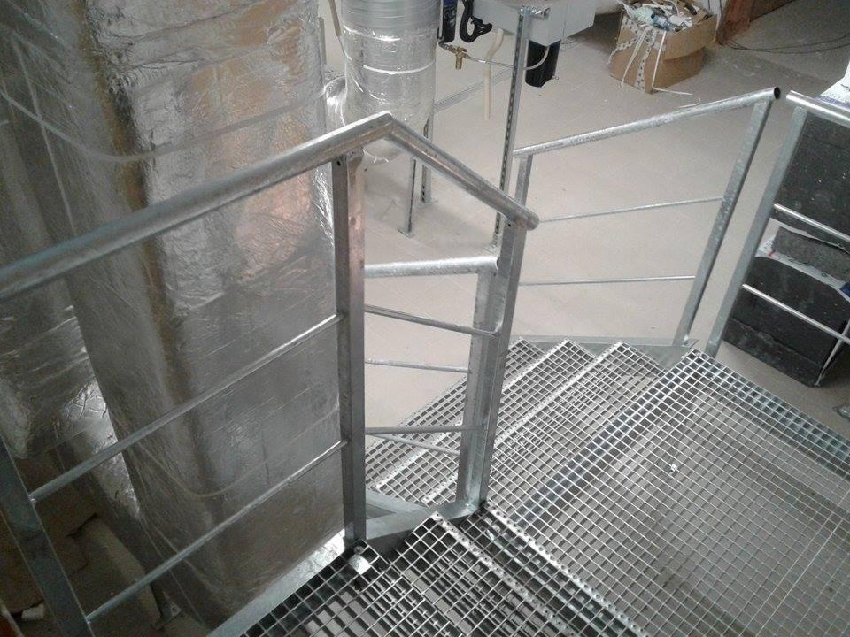 Metalowe schody z kraty pomostowej z prostą, surową balustradą z rur, widok z góry na fragment konstrukcji w pomieszczeniu technicznym.