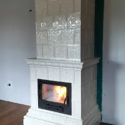 Biały, kaflowy piec kominkowy z czarnym wkładem, widoczne płomienie, dekoracyjne zdobienia na kaflach, surowe ściany w tle.