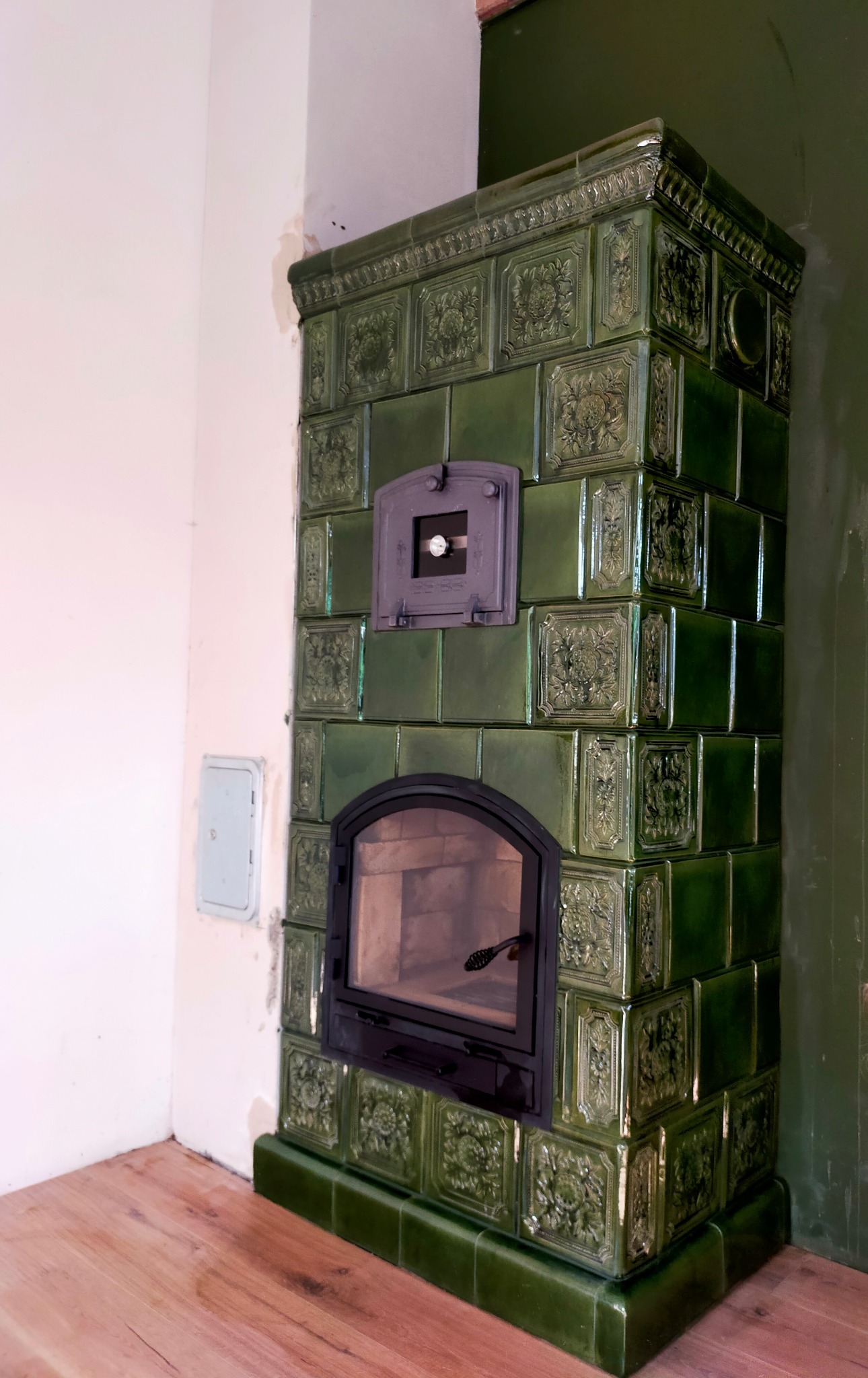 Zielony, kaflowy piec z czarną ramą drzwiczek i małym okienkiem na górze, stojący w rogu pokoju z drewnianą podłogą i białą ścianą z widoczną skrzynką elektryczną.