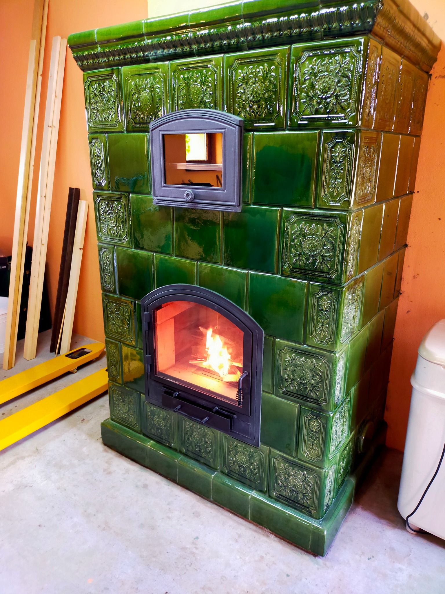 Zielony piec kaflowy z palącym się ogniem wewnątrz, widoczne dekoracyjne kafle z motywami roślinnymi, obok paleta transportowa i biały zbiornik.