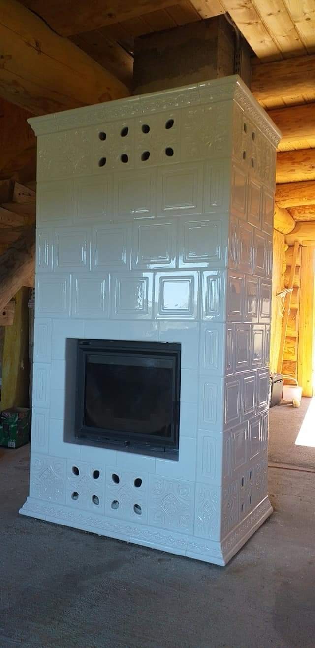 Biały piec kaflowy z czarnym wkładem kominkowym, widoczny w surowym wnętrzu drewnianego domu z bali.