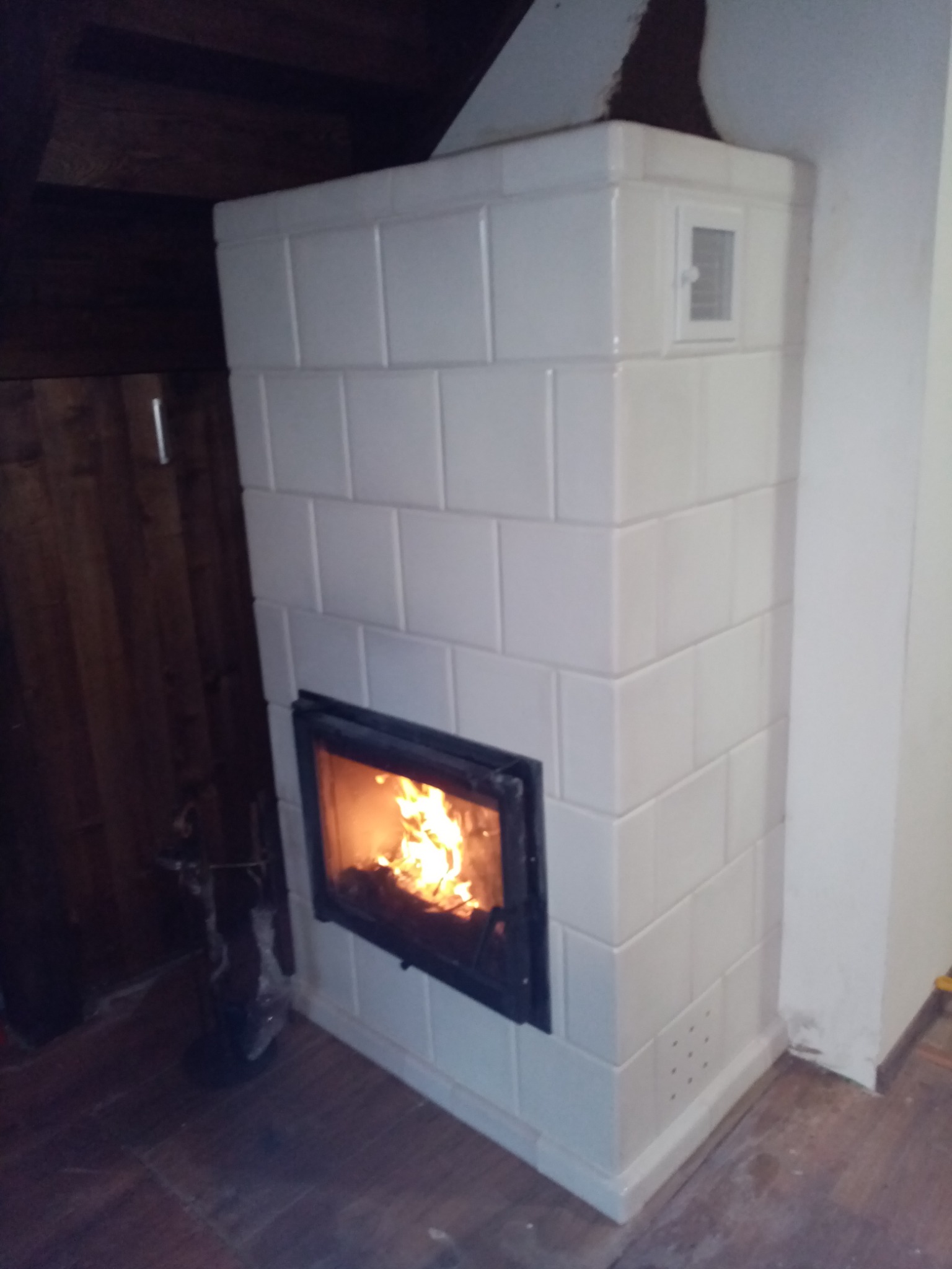 Biały piec kaflowy z palącym się ogniem w palenisku, widoczny fragment drewnianej podłogi i ściany, niewielki otwór wentylacyjny w górnej części pieca.