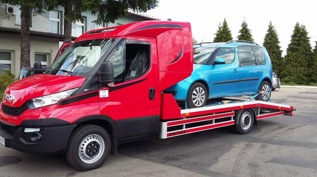 Czerwona laweta Iveco z niebieskim samochodem osobowym Skoda Roomster na platformie, zabezpieczonym pasami transportowymi, na tle budynku i drzew.
