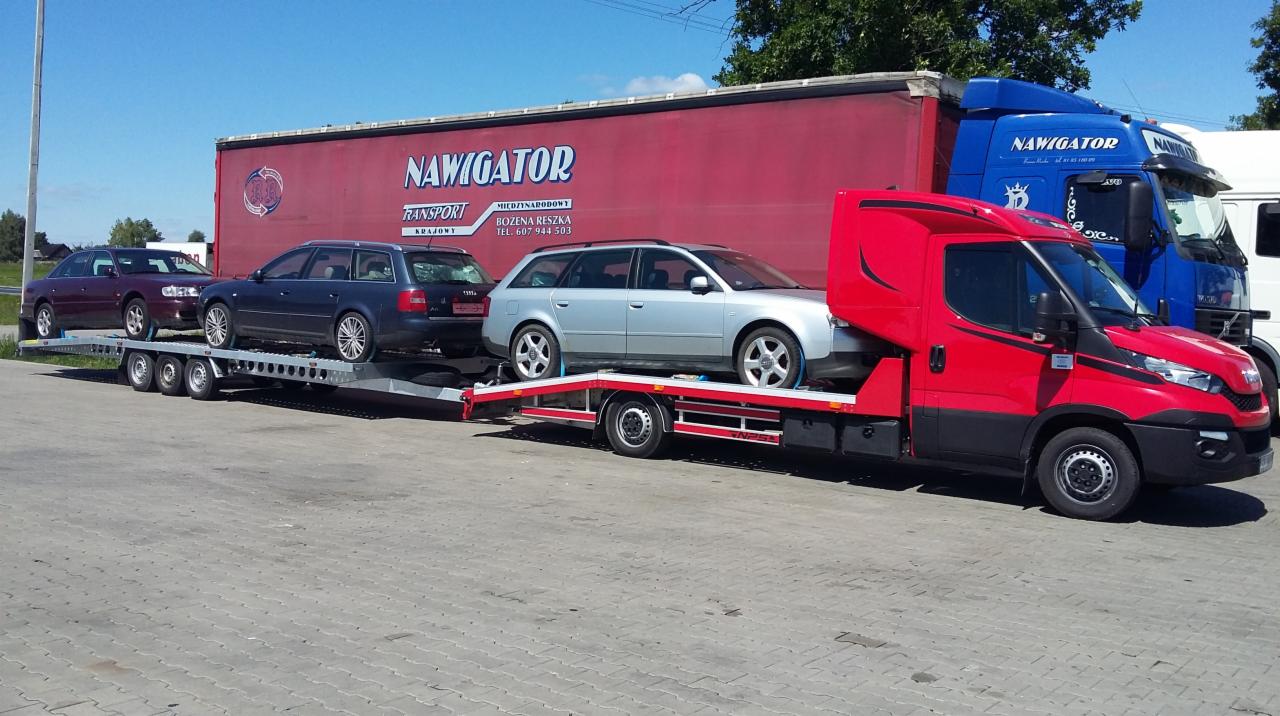 Czerwona laweta z trzema samochodami osobowymi na przyczepie, zaparkowana na betonowym placu, z widocznymi innymi ciężarówkami w tle.