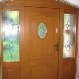 Drewniane drzwi wejściowe z szybami witrażowymi po bokach i nadświetlem łukowym, widoczna pianka montażowa.