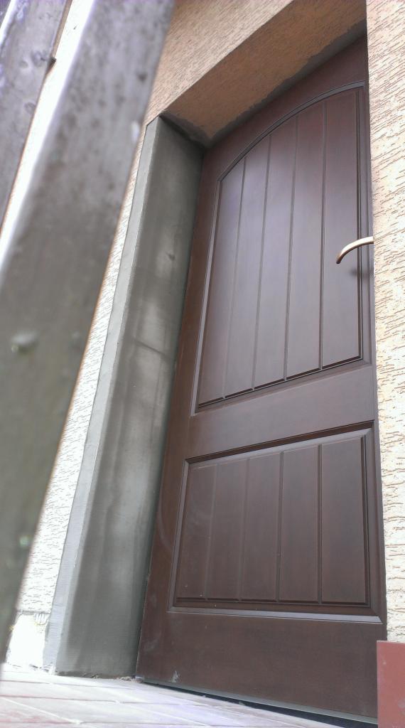 Ciemnobrązowe drzwi wejściowe z panelem i łukowatym górnym panelem, widoczne z dołu pod kątem, z fragmentem drabiny po lewej stronie i świeżo otynkowaną ościeżnicą.