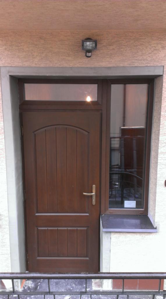 Ciemnobrązowe drzwi zewnętrzne z górnym i bocznym naświetlem, zamontowane w ościeżnicy z szarego betonu, oświetlone lampą nad wejściem.
