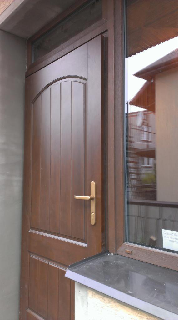 Brązowe drzwi zewnętrzne z szybą boczną, widoczny budynek odbijający się w szybie, złota klamka i okapnik.