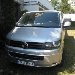 Srebrny van Volkswagen Transporter z widoczną tablicą rejestracyjną, zaparkowany na trawie przed budynkiem. Ujęcie z przodu pojazdu, w tle zieleń i zabudowania.