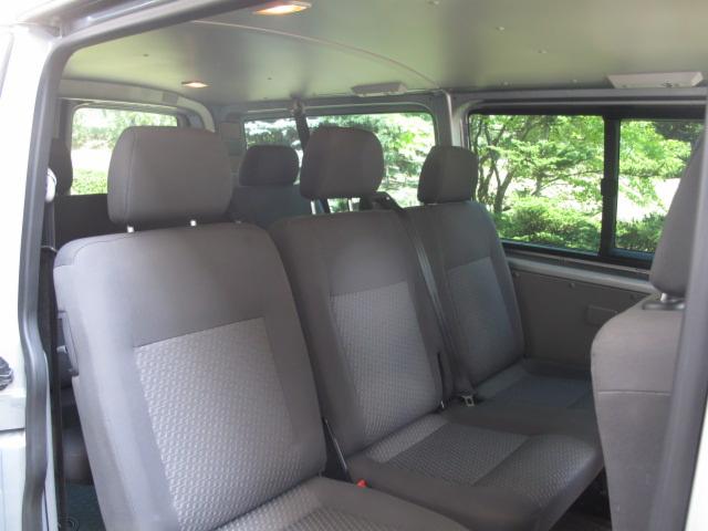 Wnętrze busa z trzema rzędami szarych foteli, widok z tyłu pojazdu, okno z widokiem na zieleń, oświetlenie sufitowe. Czyste i zadbane wnętrze pojazdu.