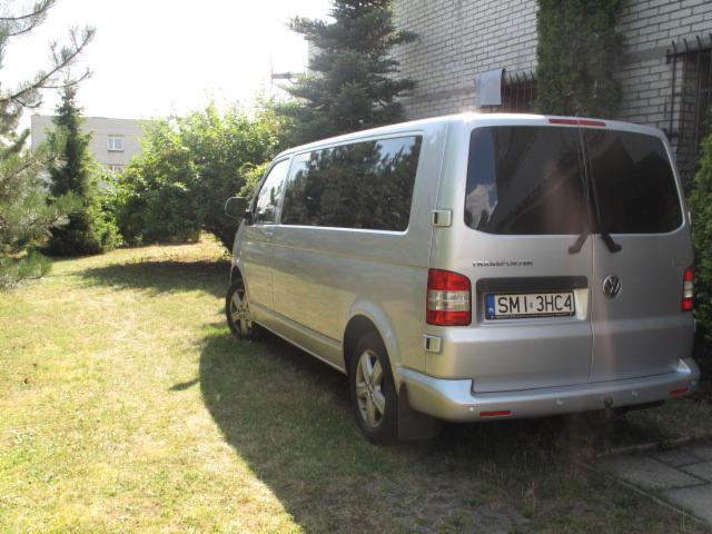 Srebrny Volkswagen Transporter zaparkowany na trawie obok budynku z cegły, z numerem rejestracyjnym SMI 3HC4. Widok z tyłu pojazdu.