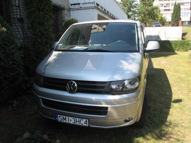 Srebrny van Volkswagen Transporter z widoczną tablicą rejestracyjną, zaparkowany na trawie przed budynkiem. Ujęcie z przodu pojazdu, w tle zieleń i zabudowania.