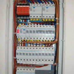 Rozdzielnica elektryczna w białej obudowie, z widocznymi zabezpieczeniami, przewodami i listwami zaciskowymi. Instalacja wykonana schludnie, z użyciem opasek.