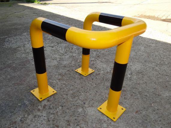 Metalowa bariera ochronna w kolorze żółto-czarnym, zabezpieczająca przed uszkodzeniami. Solidna konstrukcja na betonowym podłożu, widoczna z bliska.