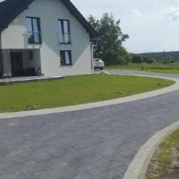 Kręty podjazd z szarej kostki brukowej prowadzący do nowoczesnego domu z białymi ścianami i ciemnym dachem, z widocznym samochodem na końcu drogi.