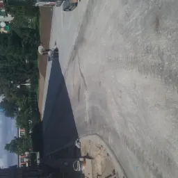 Pracownik zagęszcza walcem wibracyjnym świeżo wylaną asfaltową nawierzchnię drogi. Widoczne ślady walca na szarym betonie. W tle budynki mieszkalne i drzewa.