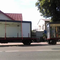 Ciężarówka z naczepą przewożąca dwie szklane konstrukcje, zabezpieczone pasami transportowymi, na tle zabudowań mieszkalnych.