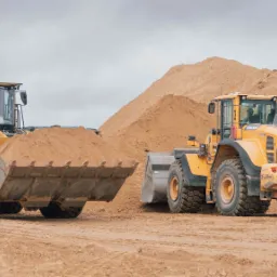 Dwa żółte wozy Volvo L180G z pełnymi łyżkami piasku na placu budowy, w tle usypana góra ziemi.