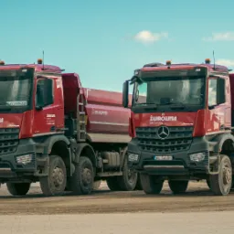 Dwa czerwone samochody ciężarowe z logo żwirowni Chojnice, zaparkowane obok siebie na placu.