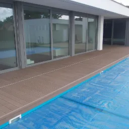 Nowoczesny taras kompozytowy w odcieniach brązu, przylegający do domu z dużymi oknami, częściowo otaczający basen przykryty niebieską folią.