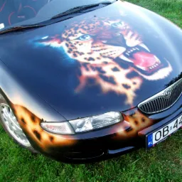 Czarny samochód Mazda z efektownym, artystycznym malunkiem przedstawiającym głowę jaguara na masce i elementami na zderzaku, stojący na trawie.