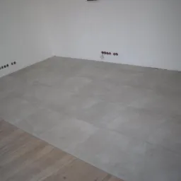 Ułożona szara terakota podłogowa w rogu pomieszczenia, z fragmentem drewnianej podłogi w lewym dolnym rogu, widoczne puszki elektryczne w ścianie.