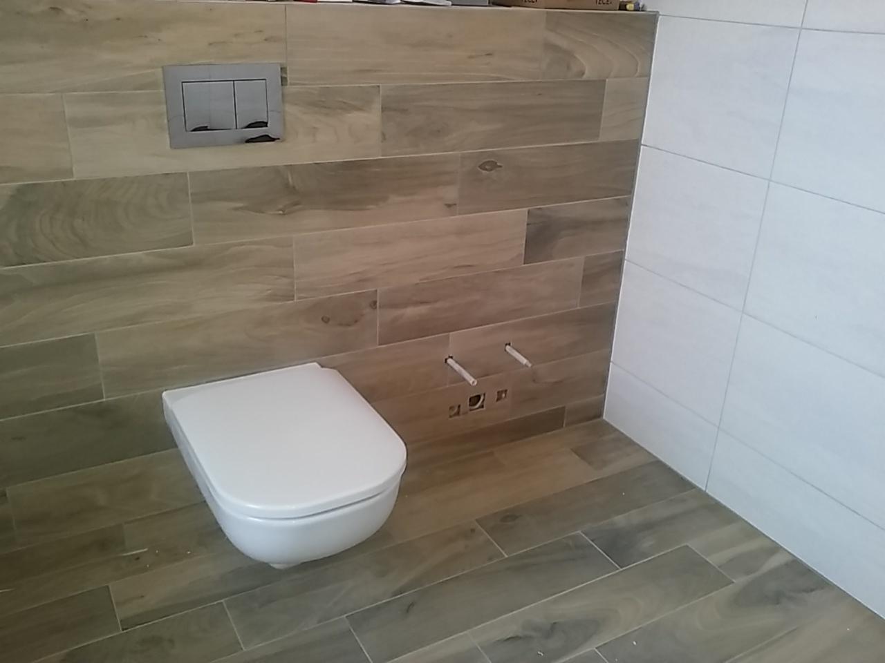 Wisząca toaleta z białym siedziskiem na tle płytek imitujących drewno i białych płytek ściennych, widoczne rury doprowadzające wodę i przycisk spłuczki.