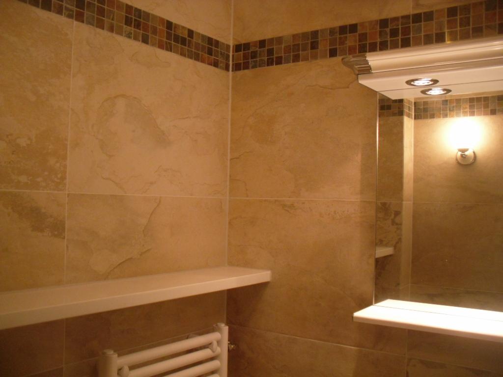 Wykończona łazienka z beżowymi płytkami imitującymi kamień, dekoracyjnym pasem mozaiki, białymi półkami i grzejnikiem drabinkowym.