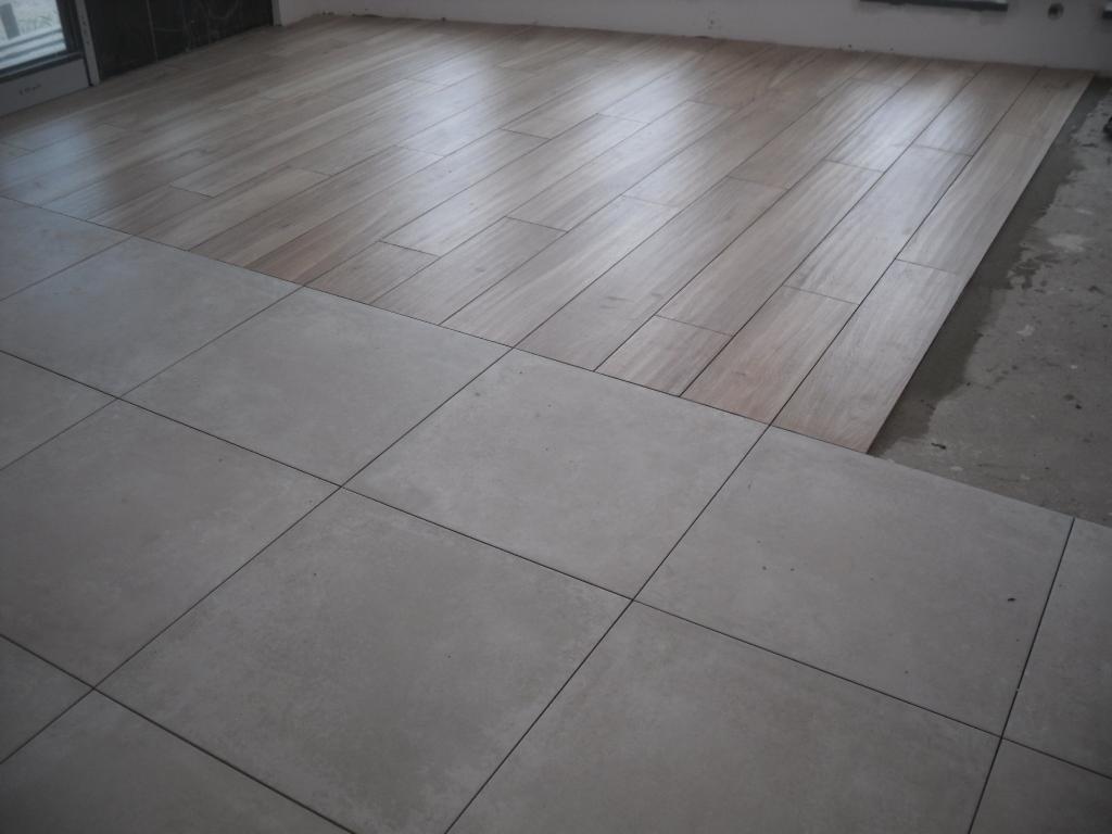 Układanie dwóch rodzajów płytek podłogowych: imitacja drewna i szare kwadratowe płytki, widoczne niedokończone krawędzie i zaprawa.