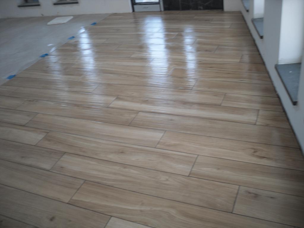 Ułożona, jasna podłoga z płytek imitujących drewno, częściowo przykryta szarym podkładem z niebieskimi taśmami zabezpieczającymi, odbijająca światło okna.