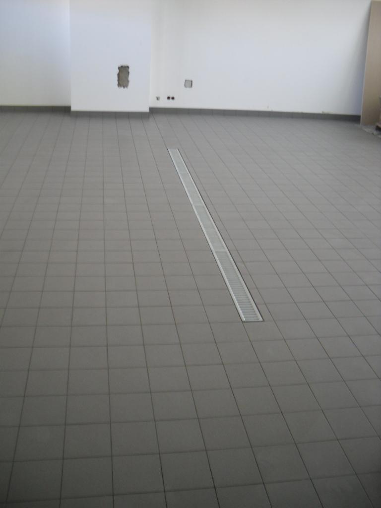 Szara podłoga wyłożona płytkami ceramicznymi z liniowym odpływem podłogowym w nowym, pustym pomieszczeniu z surowymi ścianami.