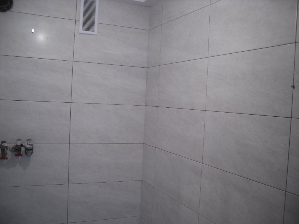 Ściana łazienki wyłożona szarymi płytkami ceramicznymi o matowym wykończeniu, widoczne zawory i kratka wentylacyjna, minimalistyczny design.