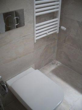 Kremowe płytki imitujące marmur w małej łazience, wisząca biała toaleta z chromowanym przyciskiem spłukiwania, drabinkowy grzejnik na ścianie.