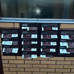 Skrzynki pocztowe z ulotkami włożonymi do większości skrzynek, umieszczone na ścianie z cegły klinkierowej pod oknem.