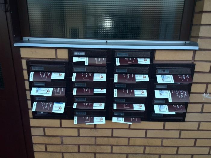 Skrzynki pocztowe z ulotkami włożonymi do większości skrzynek, umieszczone na ścianie z cegły klinkierowej pod oknem.