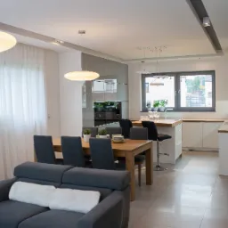 Jasne, przestronne wnętrze: salon z szarą sofą z białymi poduszkami, drewniany stół jadalny z szarymi krzesłami, kuchnia z drewnianym blatem i białymi szafkami, okno z roślinami, okrągłe lampy...